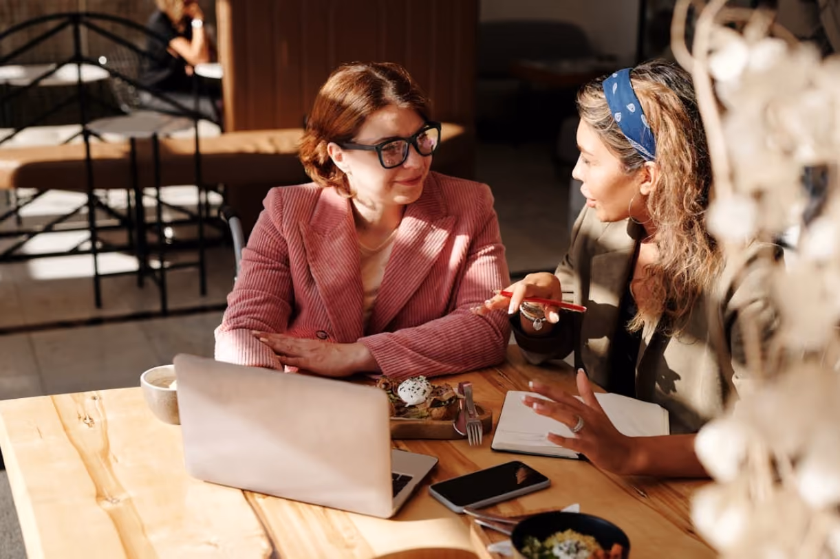 Two women sit at a sunlit café table, engaged in conversation. One wears a pink blazer, the other a blue headband. Laptops and phones are on the table.