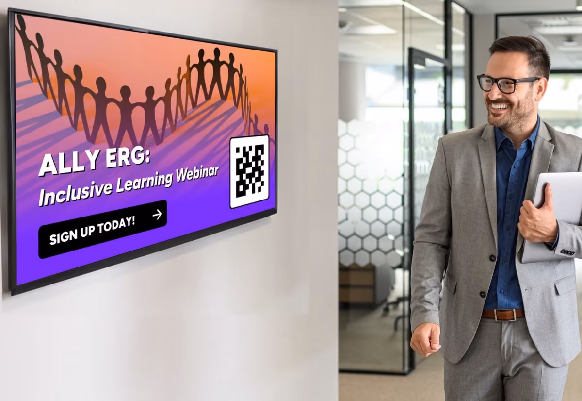 Man in a suit smiles at a wall screen showing "ALLY ERG: Inclusive Learning Webinar" with a QR code and "SIGN UP TODAY!" button, in a modern office.