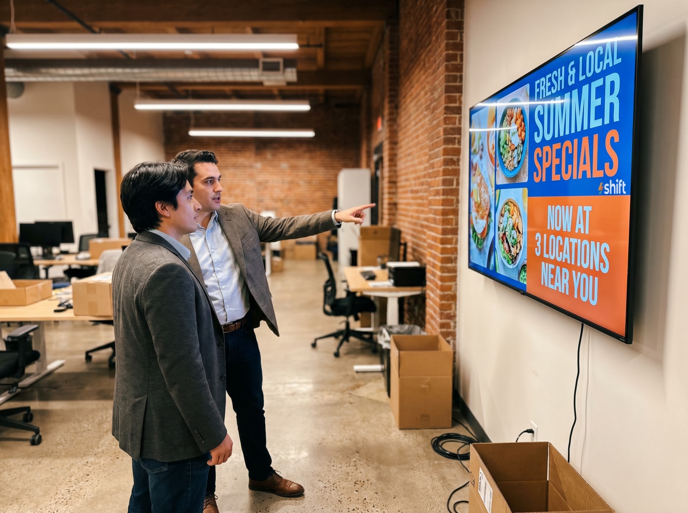 Two men in an office point at a screen displaying "Fresh & Local Summer Specials" with vibrant food images and text.