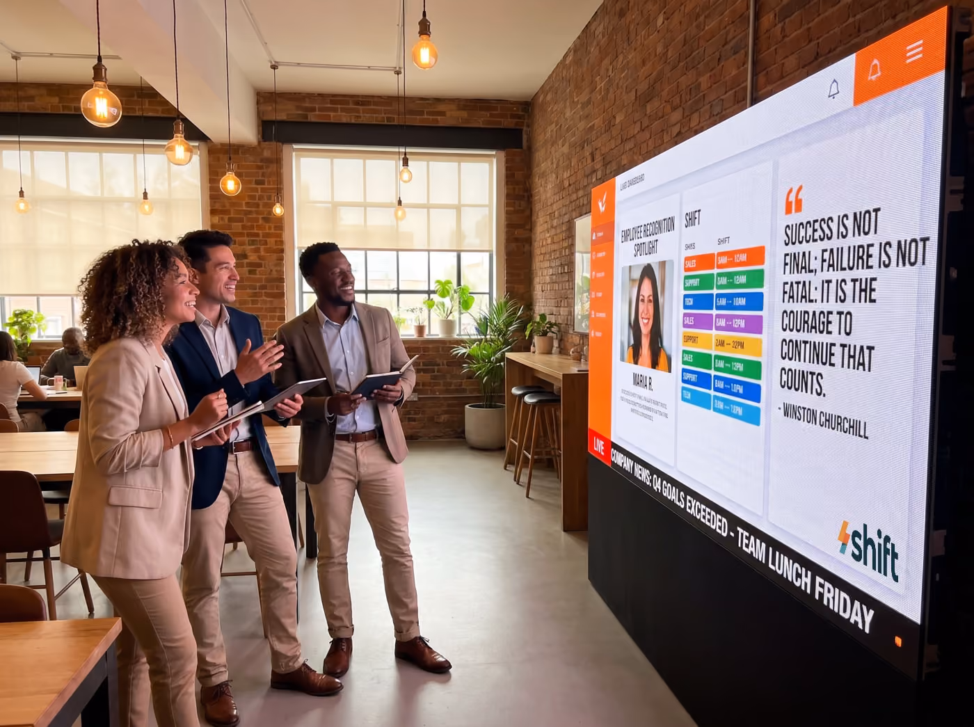 Three colleagues in a modern office smile at a large screen displaying employee recognition and motivational quotes.