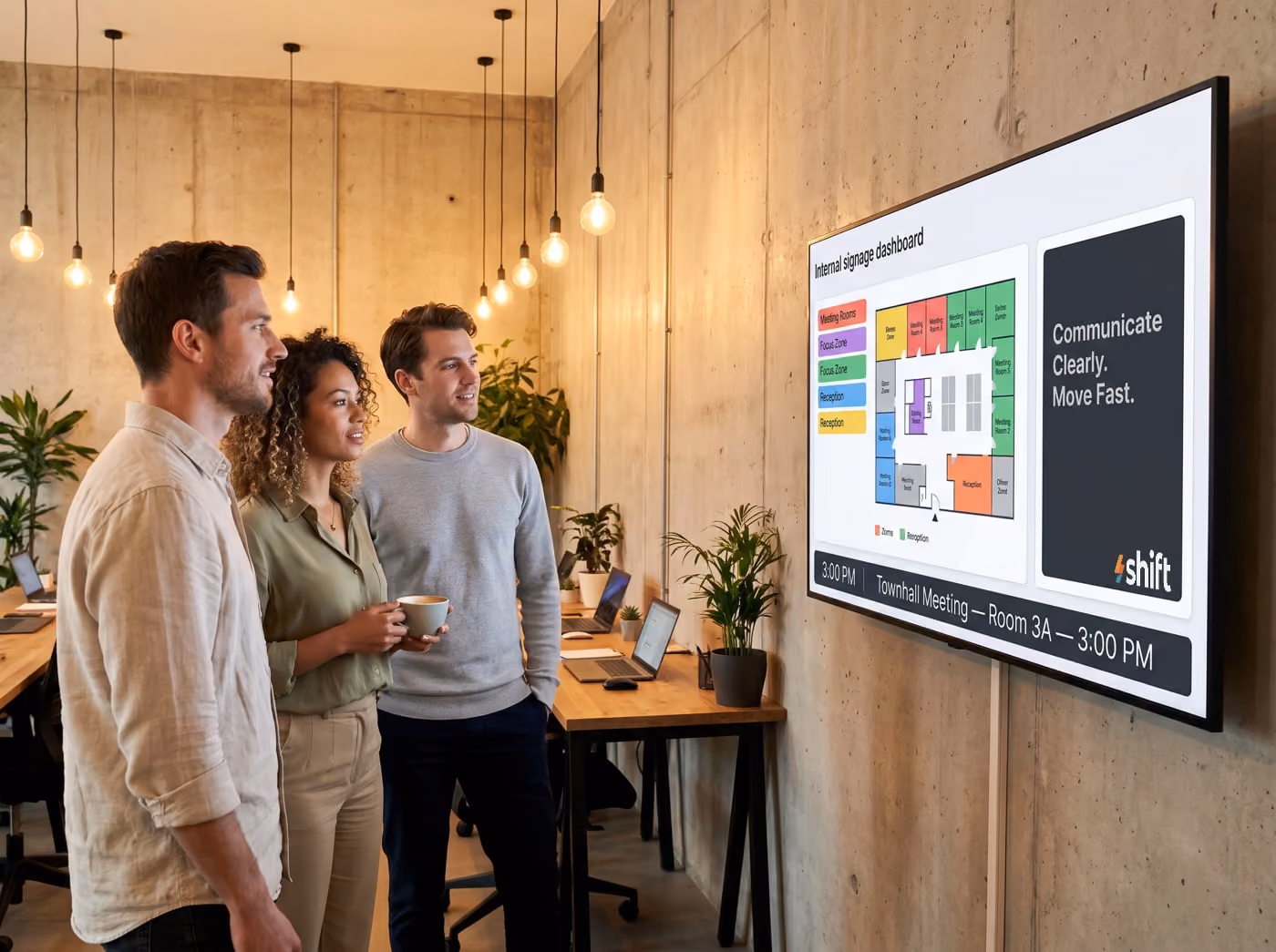 Three colleagues in a modern office discuss a digital dashboard displaying meeting room info and the message "Communicate Clearly.