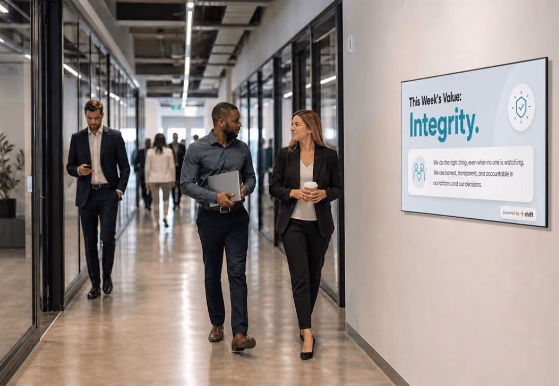 Two colleagues walk past a sign about integrity in a modern office hallway, engaging in conversation and holding a laptop and coffee.