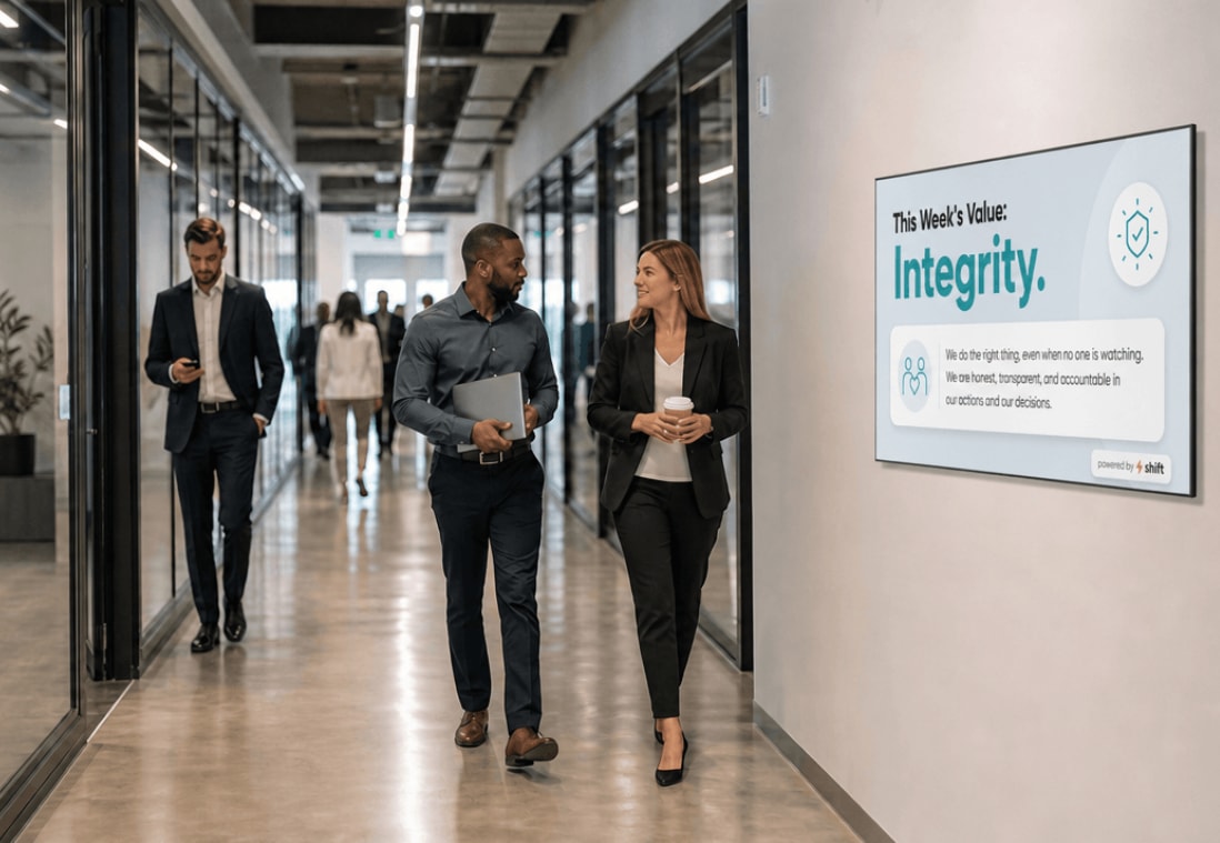 Two colleagues walk past a sign about integrity in a modern office hallway, engaging in conversation and holding a laptop and coffee.