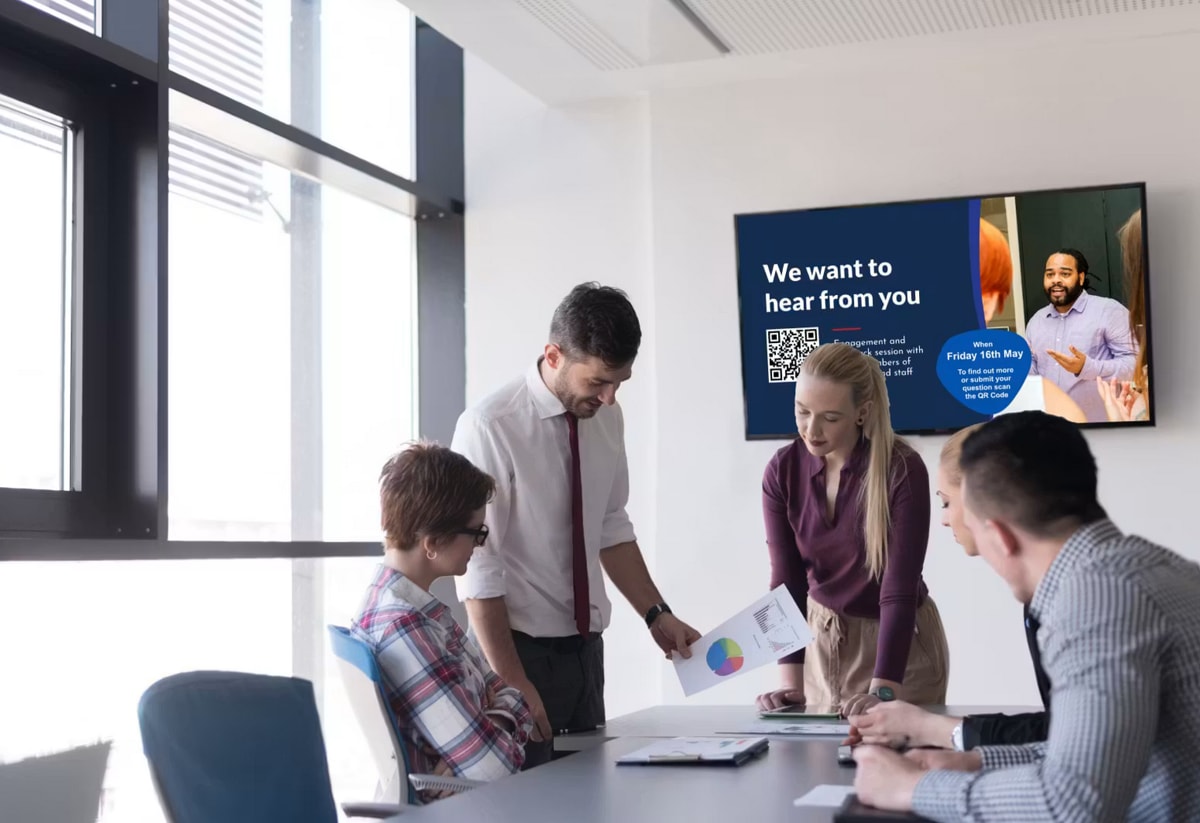 A team discusses a chart in a meeting room. A screen displays "We want to hear from you" with a QR code and event details.