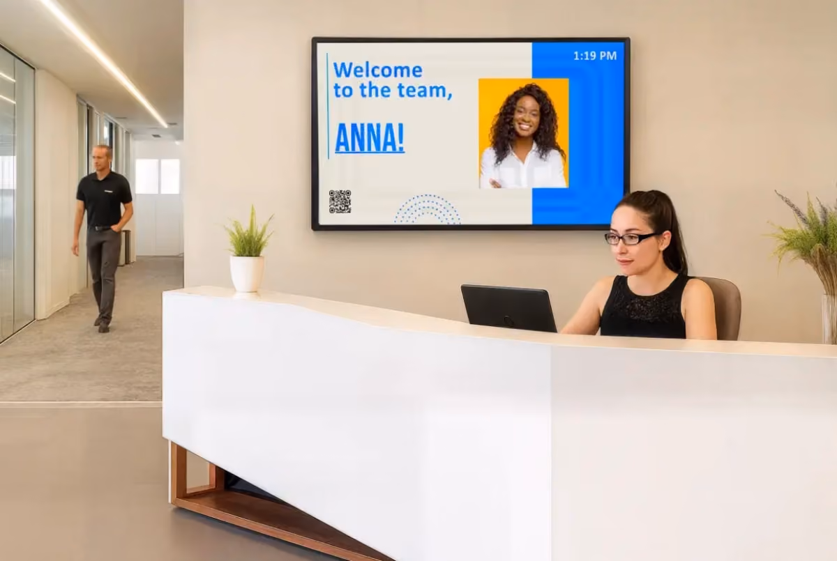 Receptionist at desk with laptop, welcome sign for Anna on screen, man walking in hallway, plants on counter, modern office.