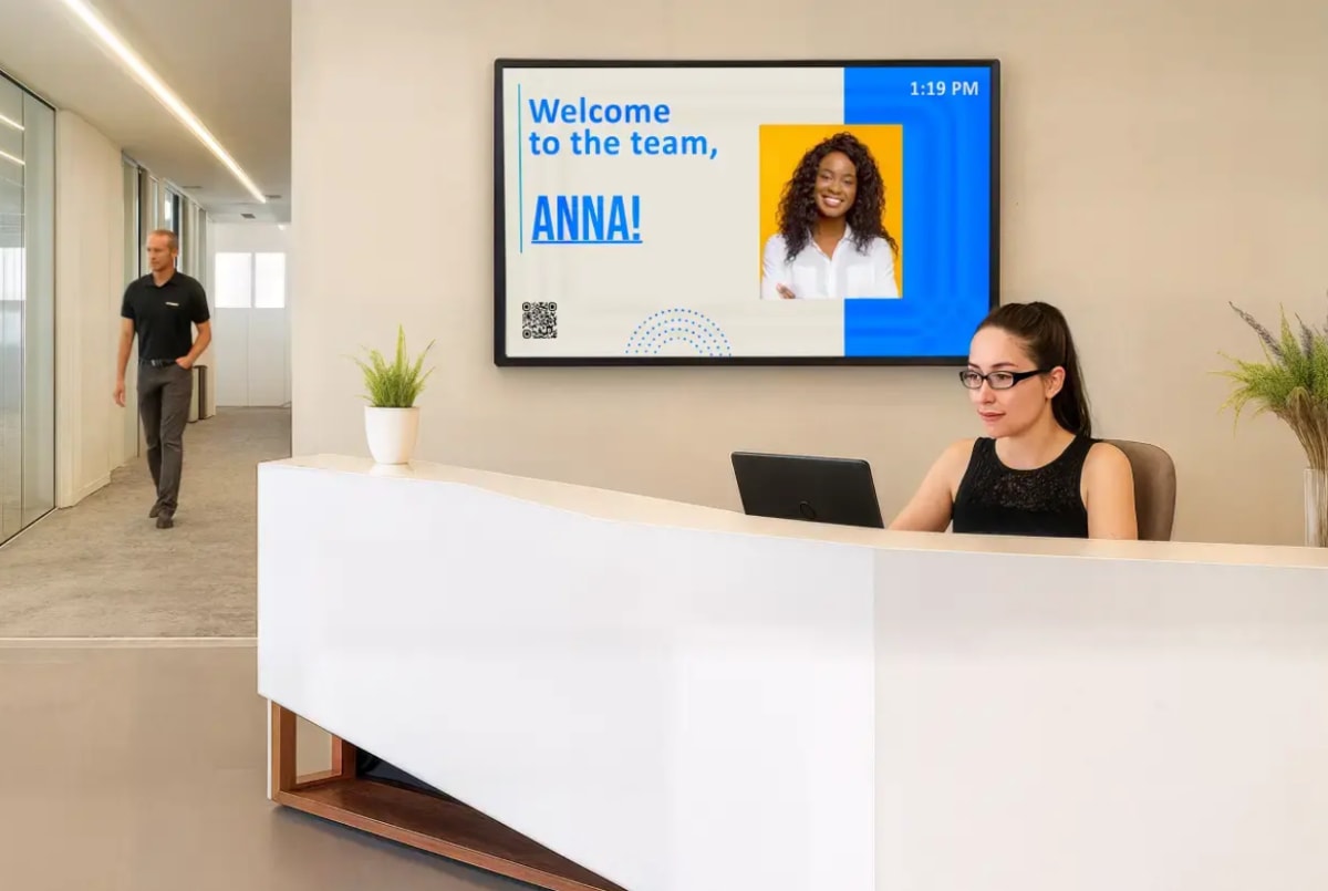 Receptionist at desk with laptop, welcome sign for Anna on screen, man walking in hallway, plants on counter, modern office.