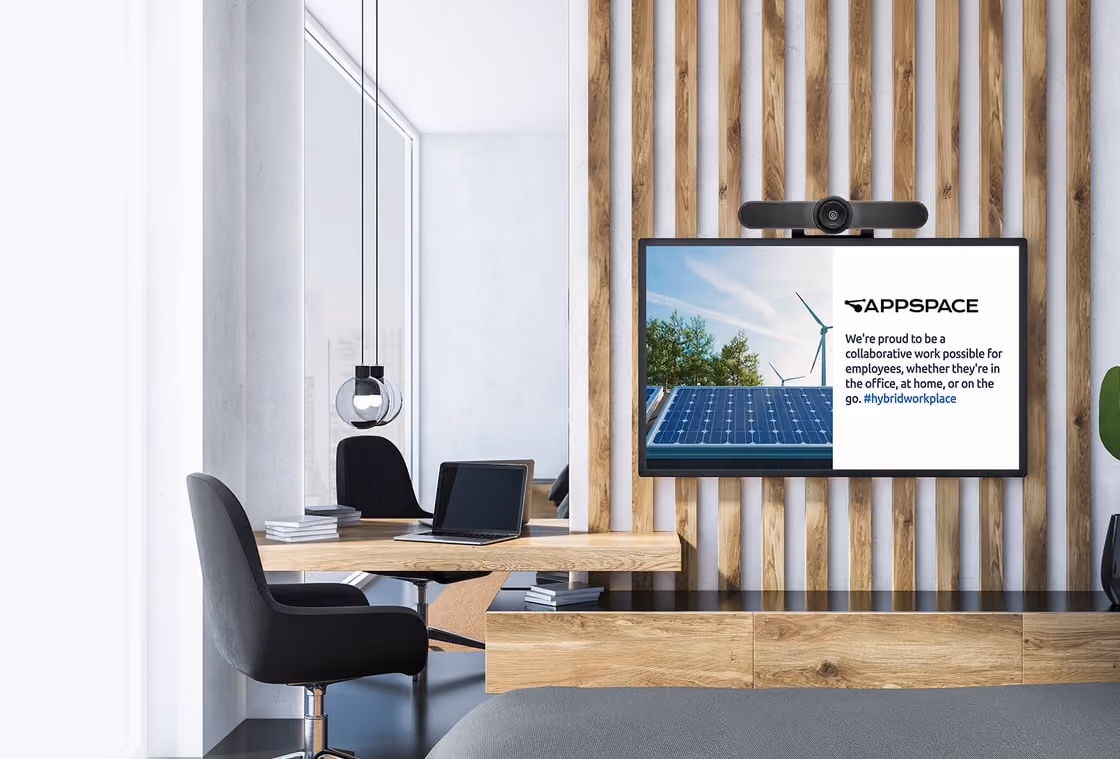Modern office with a wooden desk, black chair, and screen displaying Appspace message about hybrid workplace collaboration.