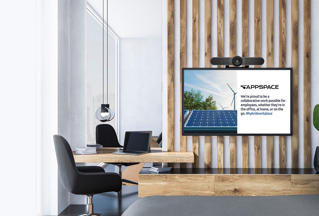 Modern office with a wooden desk, black chair, and screen displaying Appspace message about hybrid workplace collaboration.