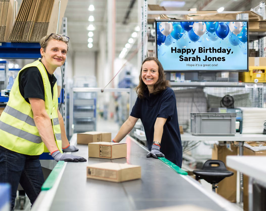 Two individuals stand in a warehouse beside a conveyor belt loaded with boxes, overseeing the sorting process.