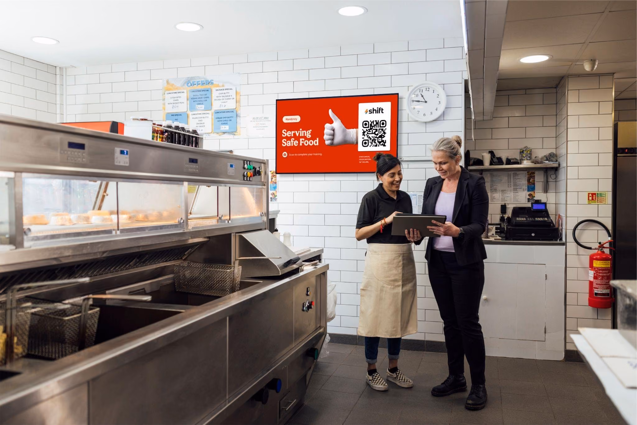 Two women stand in a kitchen, with a QR code displayed on the wall behind them.