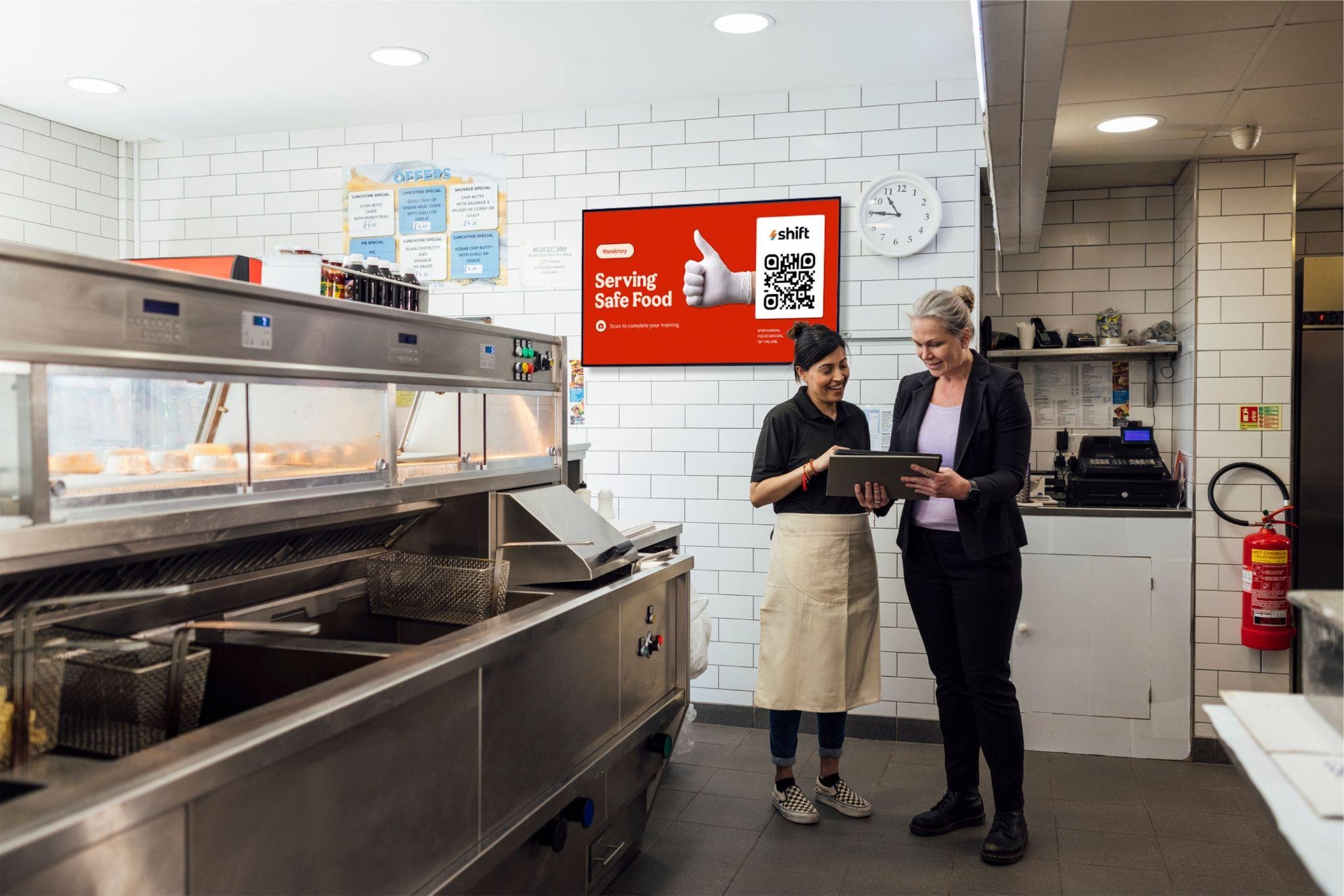 Two women stand in a kitchen, with a QR code displayed on the wall behind them.