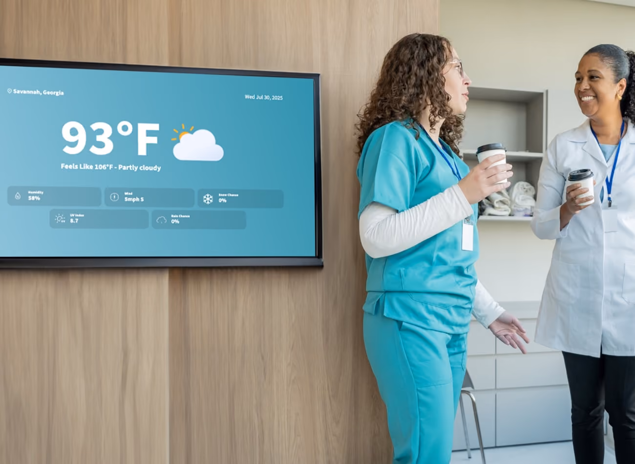 Two women in scrub suits stand together in front of a television, engaged in conversation or viewing content.