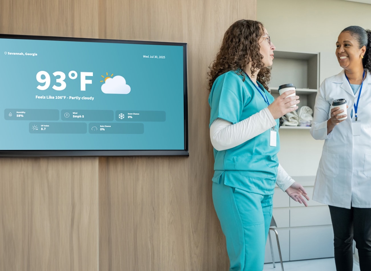 Two women in scrub suits stand together in front of a television, engaged in conversation or viewing content.