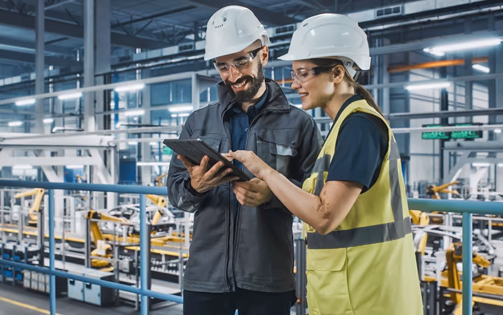 Two individuals wearing hard hats stand in front of a factory, showcasing a safety-conscious work environment.