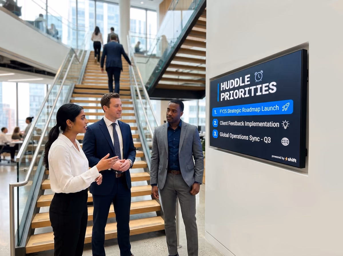 Three professionals discuss "Huddle Priorities" on a screen in a modern office with a staircase and people in the background.