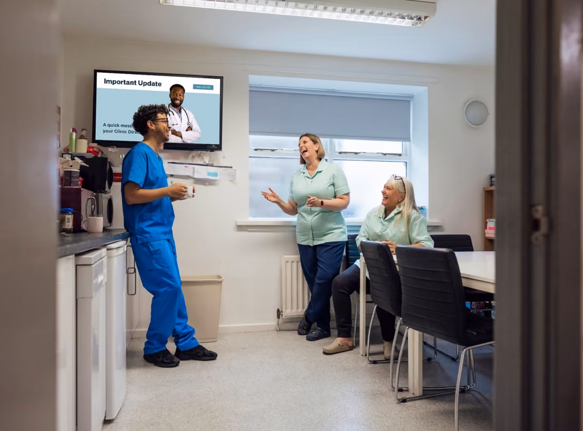 Three healthcare workers in scrubs chat in a break room, with a wellness poster on the wall promoting well-being resources.