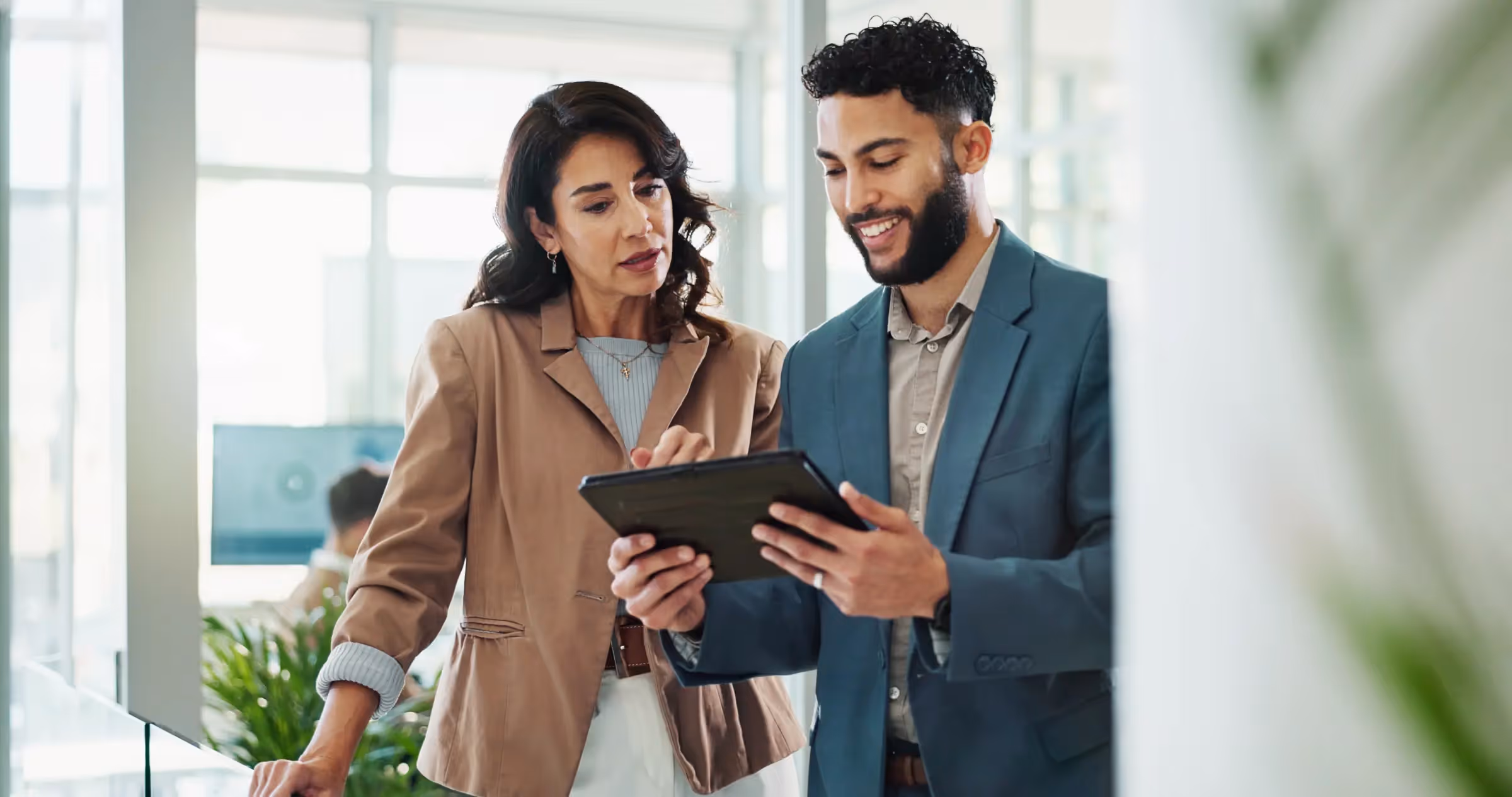 Two business colleagues, a woman and a man, discussing something while looking at a tablet in a bright office.
