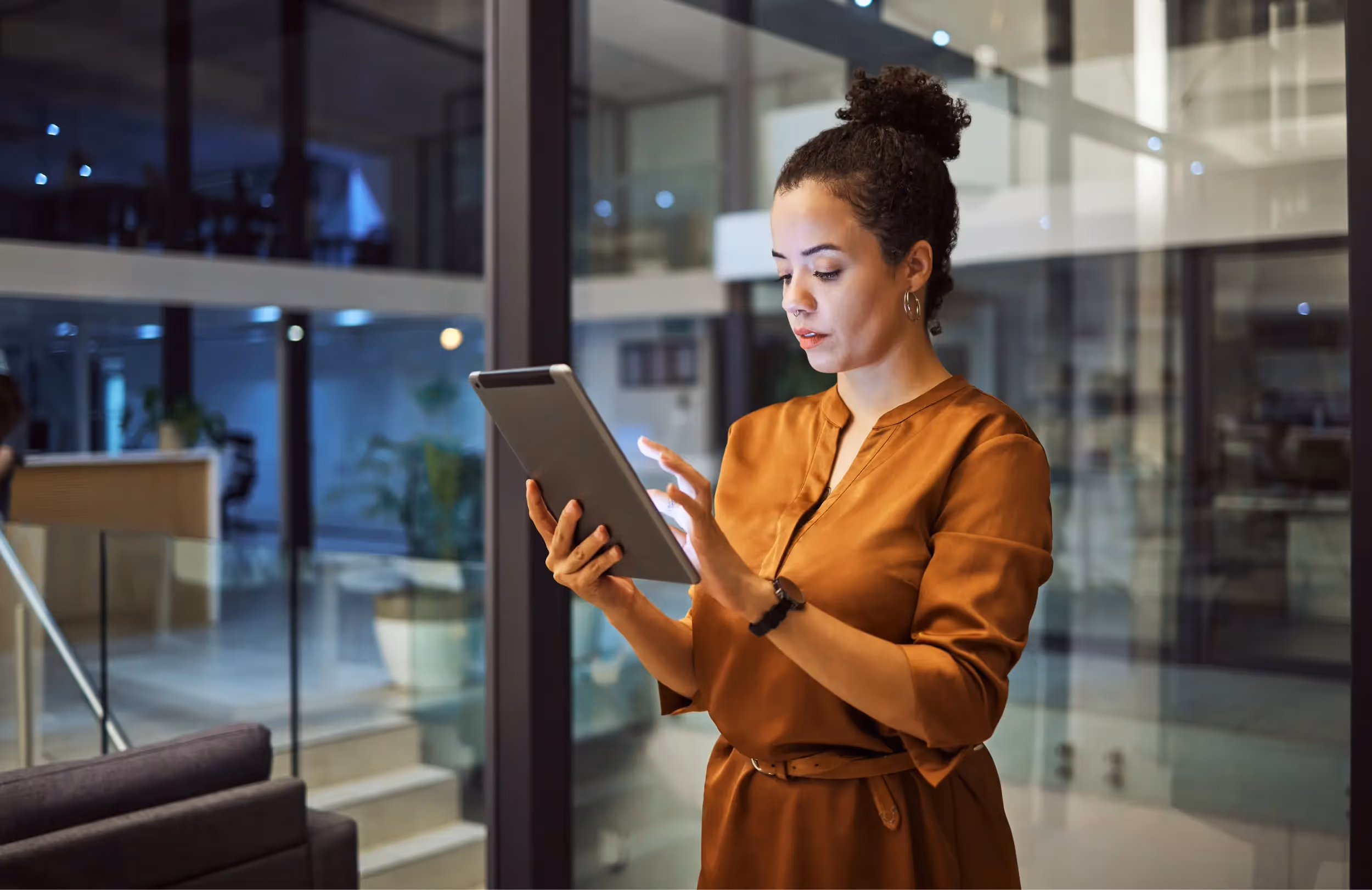 Woman in a brown dress using a digital tablet in a modern office with glass walls.