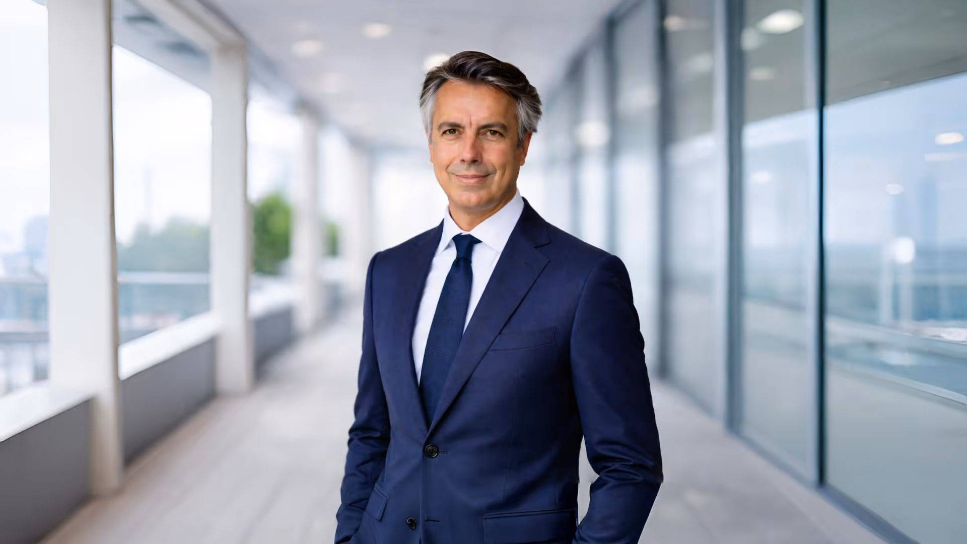 Confident middle-aged man in a navy blue suit and tie standing in a modern glass-walled corridor.