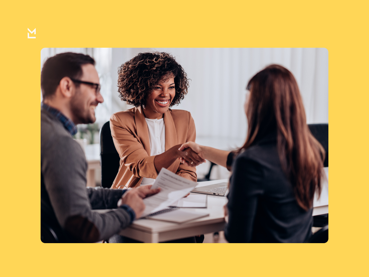 Three professionals in an office setting, with two shaking hands across a table and one smiling while holding documents, suggesting a successful meeting or agreement.
