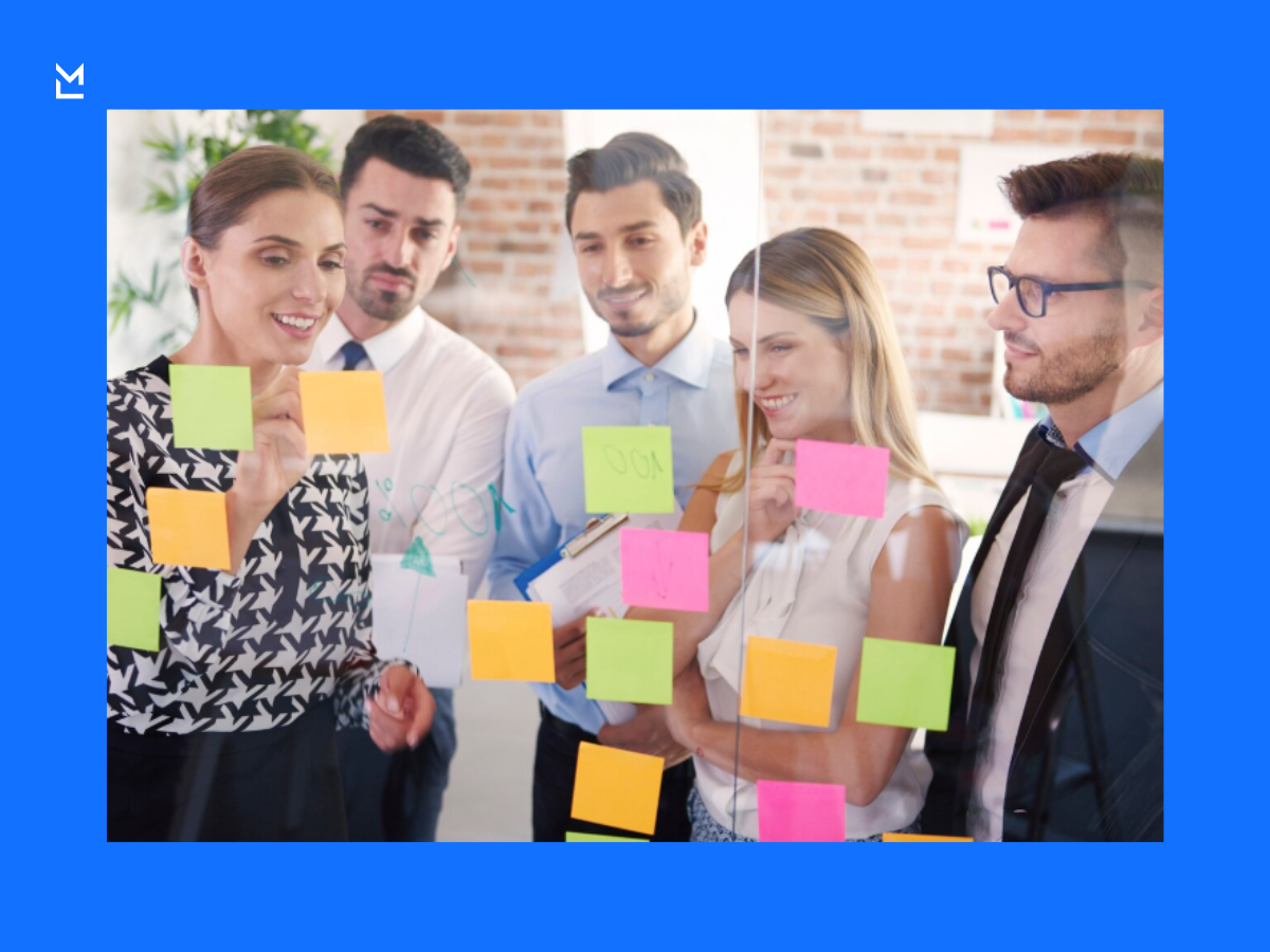 Five people brainstorming at a glass wall covered with colorful sticky notes, collaboratively planning and prioritizing ideas.
