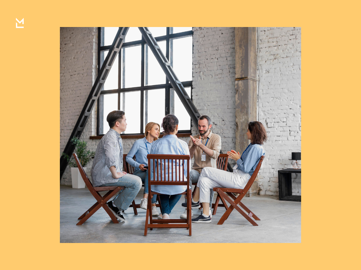 Group of five people seated in a circle in an industrial-style room, engaged in a discussion with one person speaking and others listening attentively.