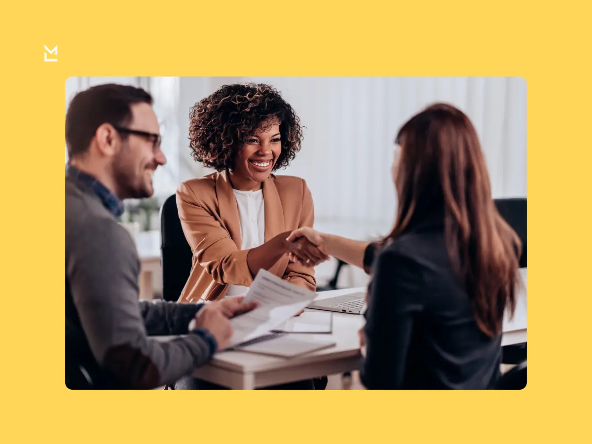 Three professionals in an office setting, with two shaking hands across a table and one smiling while holding documents, suggesting a successful meeting or agreement.