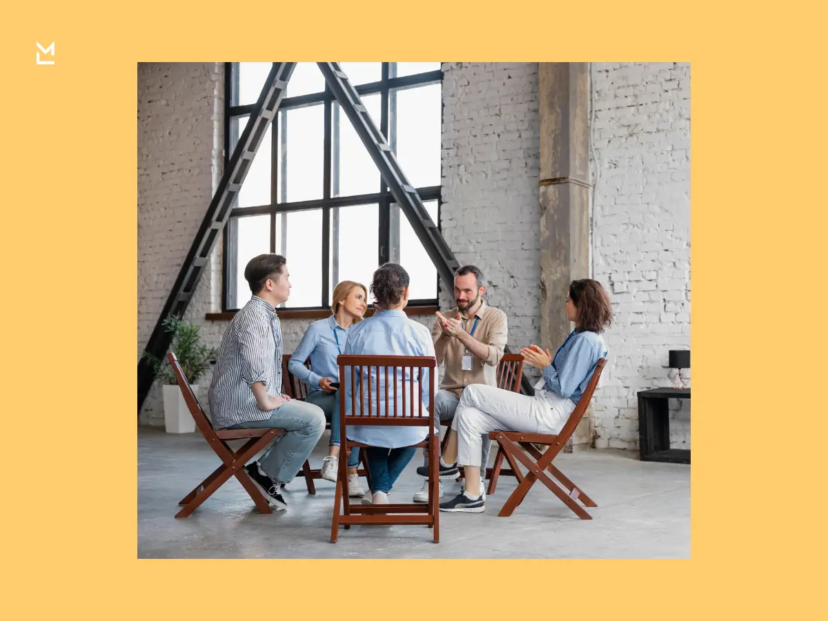 Group of five people seated in a circle in an industrial-style room, engaged in a discussion with one person speaking and others listening attentively.
