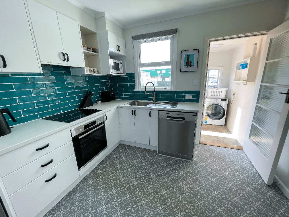 Accommodation in Gisborne Flat 4, 2 bedroom holiday house new kitchen with white cabinets, freestanding fridge, oven, microwave and dishwasher, vinyl tile flooring window and adjoining laundry area with washing machine