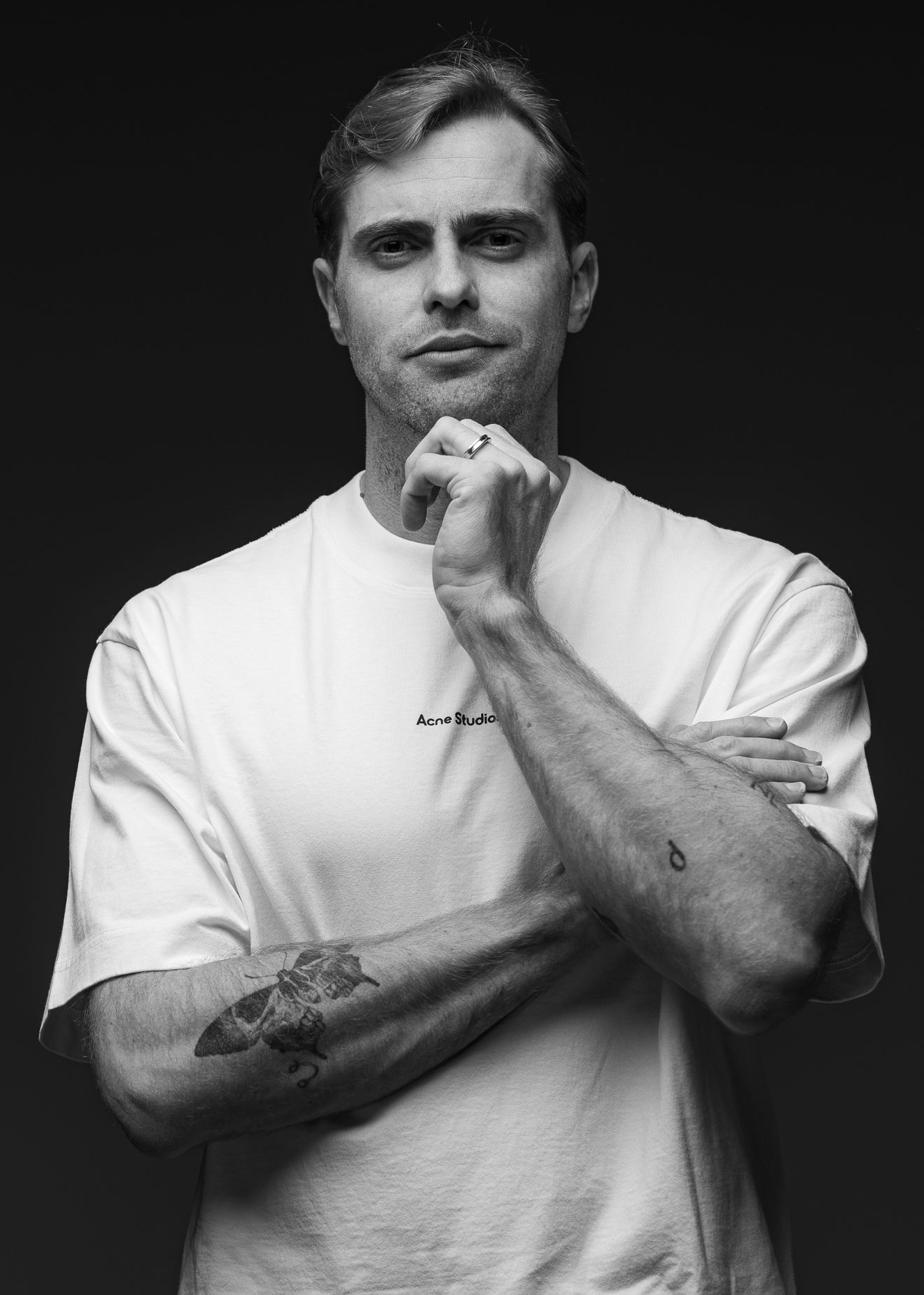 Thirty something man wearing white shirt, looks confidently ahead. Tattoos adorn his forearms. 