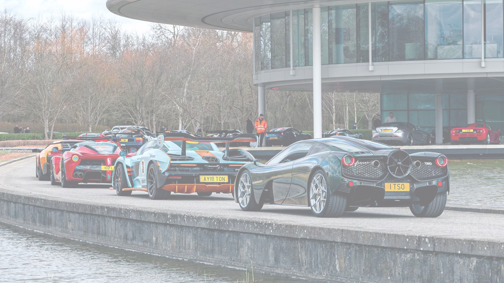 Line of luxury sports cars including McLaren Senna, Ferrari LaFerrari, and Pagani Huayra parked near modern glass building beside water.