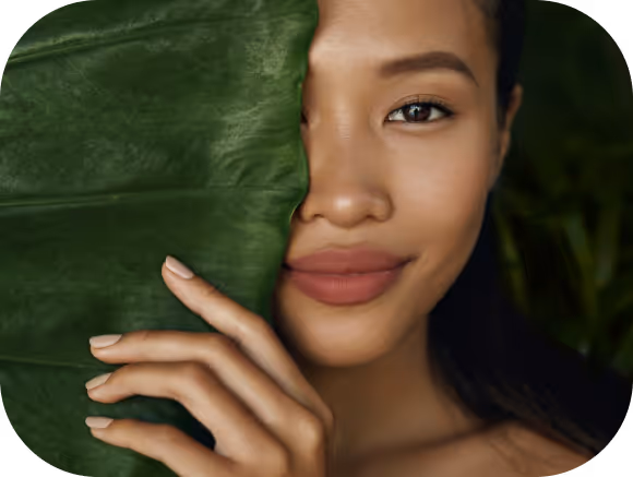 Close-up of a woman with smooth skin partially covering her face with a large green leaf.