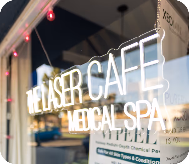 Illuminated sign reading 'The Laser Cafe Medical Spa' reflected on a glass window with blurred street background.