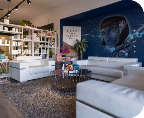 Modern waiting area with white leather sofas, a round reflective coffee table with skincare products, a shaggy gray rug, and a wall mural of a woman with dark hair.