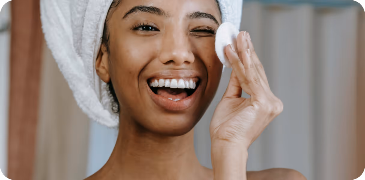 Smiling woman with a white towel wrapped around her head gently cleansing her face with a cotton pad.