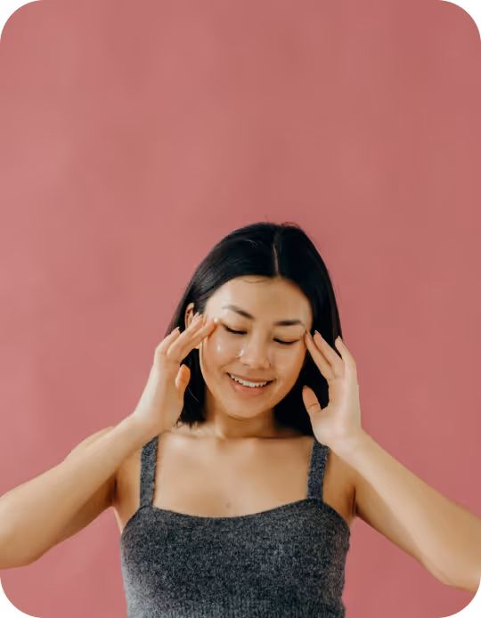 Smiling young woman with under-eye gel patches gently touching her temples against a pink background.