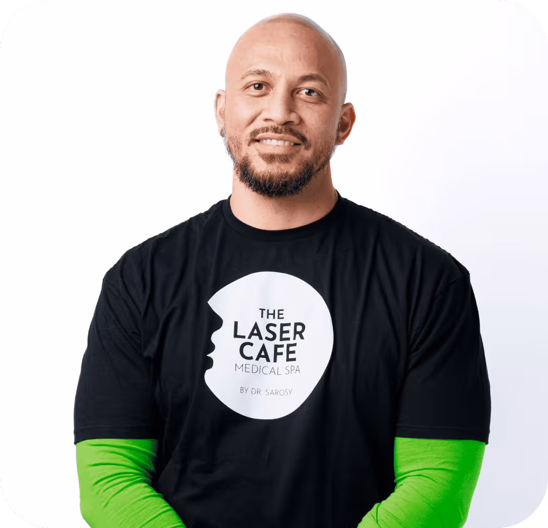 Smiling bald man with a beard wearing a black t-shirt over a bright green long-sleeve shirt, featuring The Laser Cafe Medical Spa logo.
