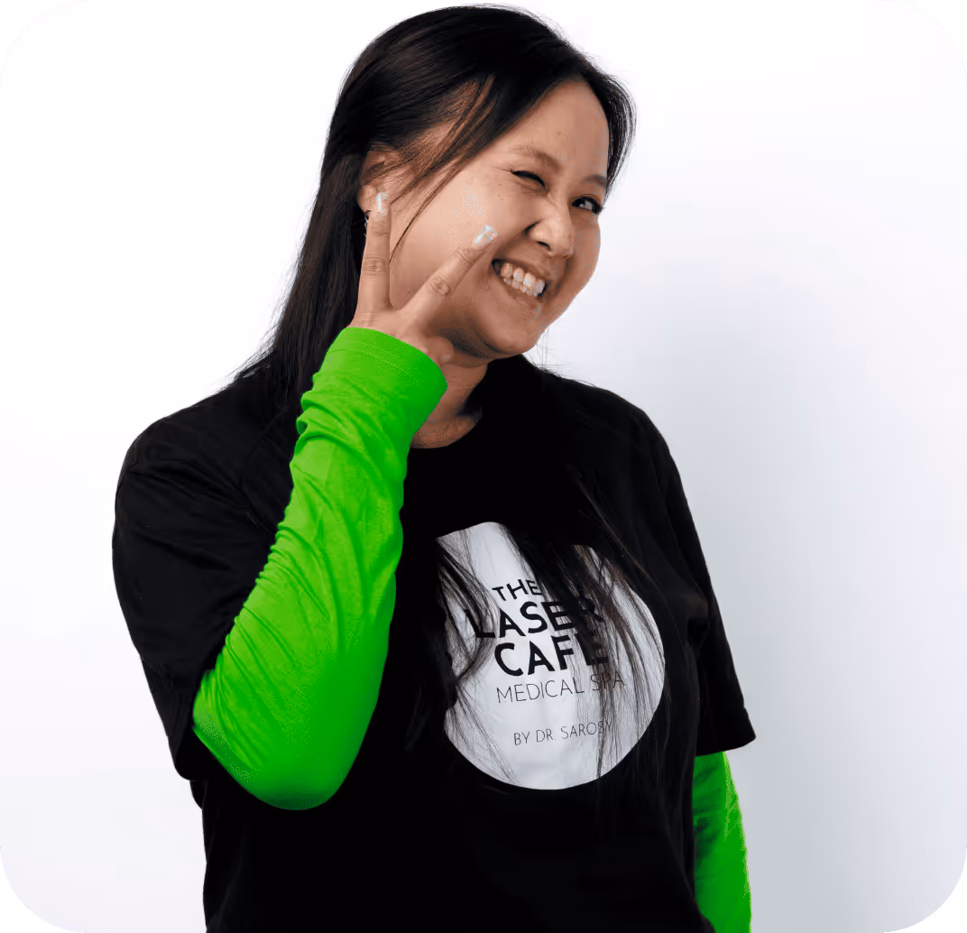 Smiling woman in a black The Laser Cafe Medical Spa t-shirt and bright green long sleeves making a peace sign and winking.