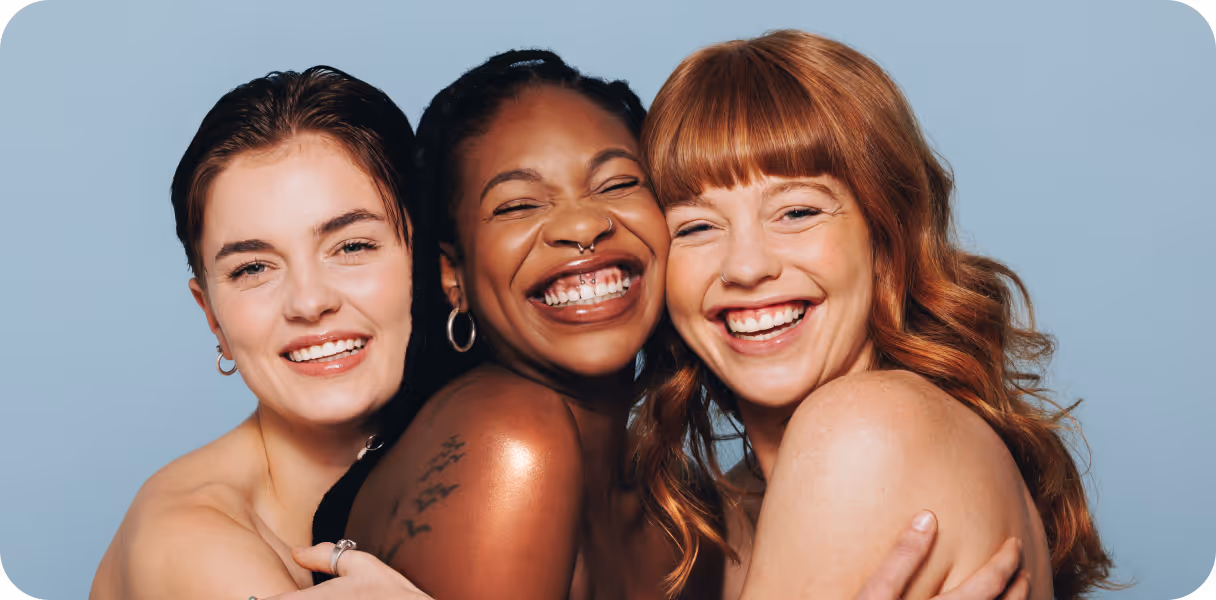 Three smiling women with different skin tones and hairstyles embracing each other against a blue background.