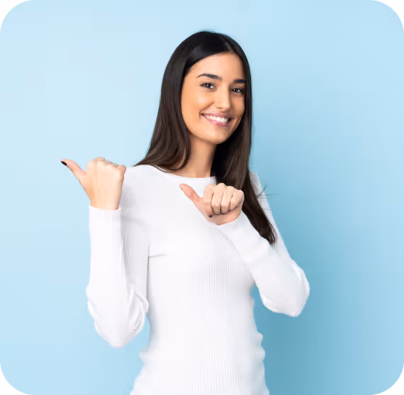 Smiling young woman in white sweater pointing thumbs back over her shoulders against a light blue background.