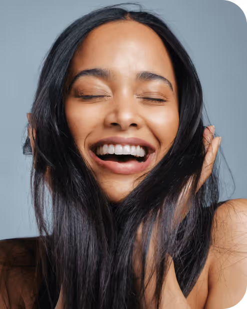 Close-up of a woman with long dark hair smiling with eyes closed, holding her face with both hands against a plain background.