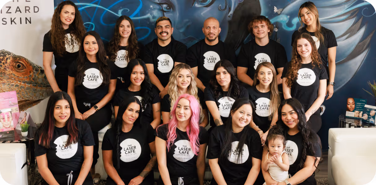 Group of diverse people wearing black The Laser Cafe Medical Spa T-shirts posing indoors against a colorful mural background, including one woman holding a baby.