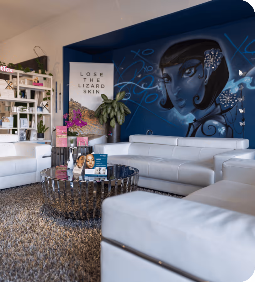 Modern waiting area with white leather sofas, a reflective coffee table displaying skincare brochures, a large plant, and a blue wall mural of a stylized woman’s face.