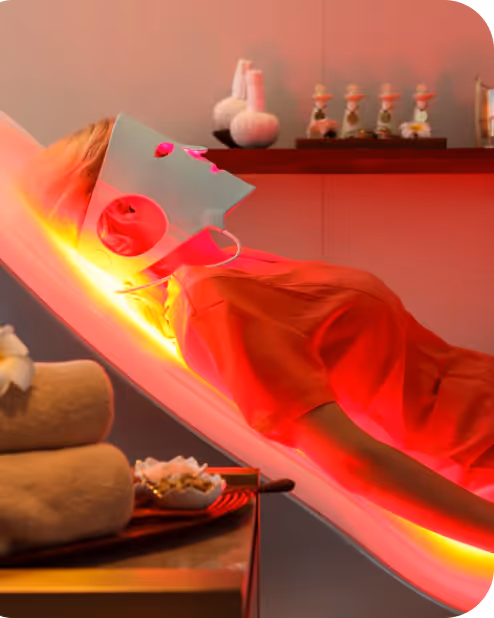 Person lying on a reclined chair wearing a futuristic white LED light therapy face mask emitting red and yellow light.