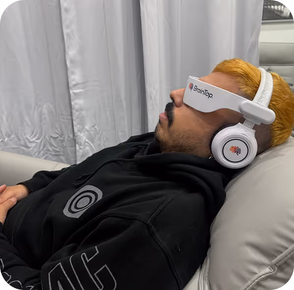Person with blond hair lying back on a couch wearing white BrainTap headphones and a black hoodie.