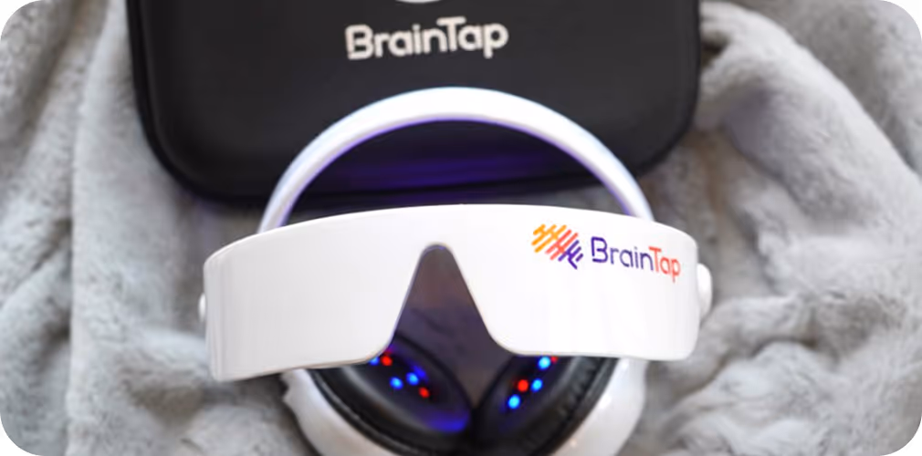 White BrainTap headset with glowing blue and red lights resting on a soft gray blanket alongside a black BrainTap carrying case.
