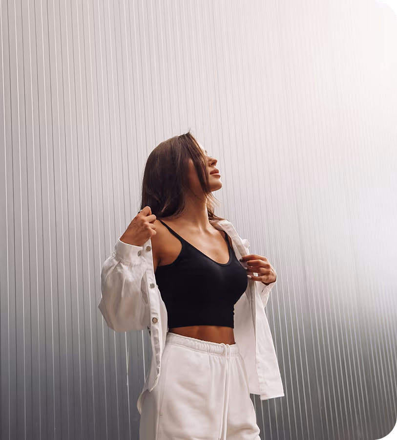 Woman looking up wearing a black crop top, white jacket, and white sweatpants against a ribbed metal background.