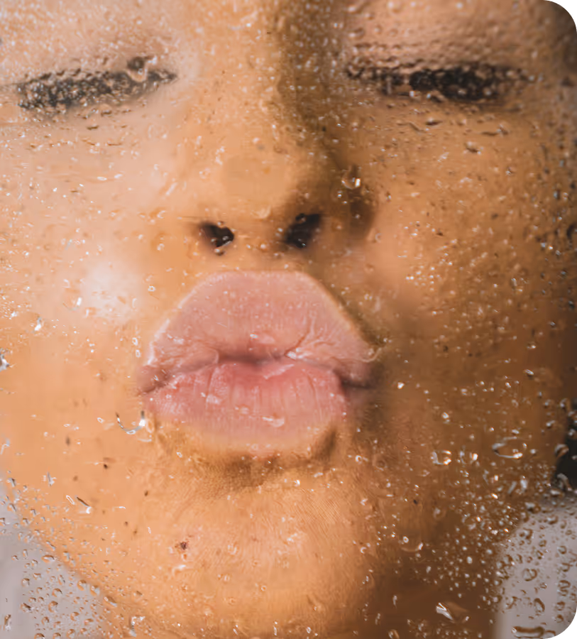 Close-up of a person's face with eyes closed and lips puckered behind a water-covered glass surface.