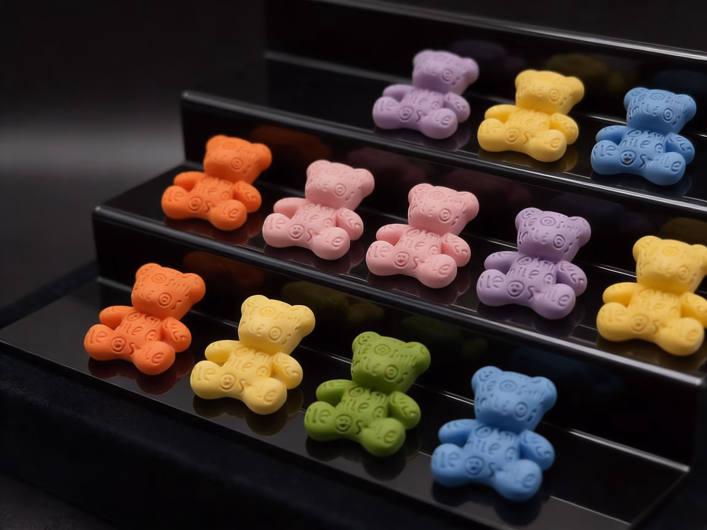 Colorful bear-shaped nail charms with smiley text arranged on glossy black display steps.