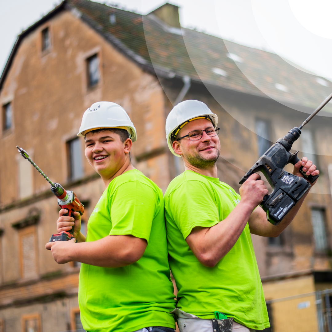Zwei Menschen in passenden grünen Hemden umarmen sich glücklich in einer belebten Inneneinrichtung, umgeben von anderen, die angeregt über ein spannendes Bauvorhaben diskutieren.